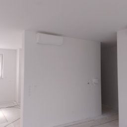 Wykończenia wnętrz - Białe wnętrze po remoncie. Widoczne gładkie ściany, zakryty klimatyzator, przygotowane podłoże pod panele. Minimalistyczny design, jasne oświetlenie, świeżo po pracach.