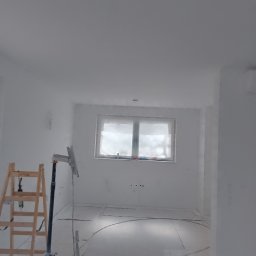 Wykończenia wnętrz - Białe, świeżo pomalowane wnętrze z drabiną i osłoniętym oknem. Widoczny sprzęt malarski sugeruje prace wykończeniowe w trakcie realizacji.