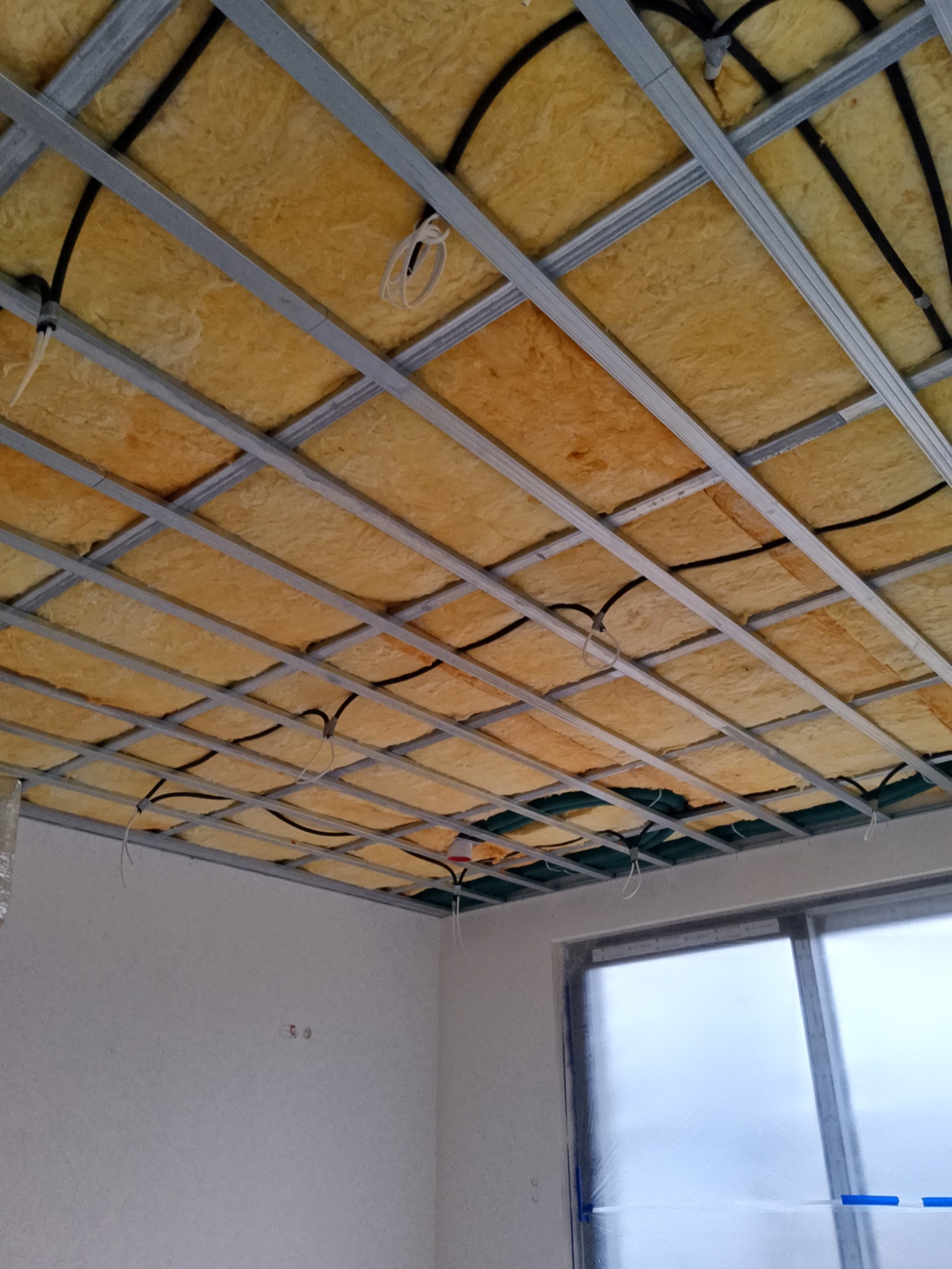Metalowa konstrukcja sufitu podwieszanego z izolacją z wełny mineralnej i przewodami elektrycznymi, widok z dołu, okno zabezpieczone folią malarską.