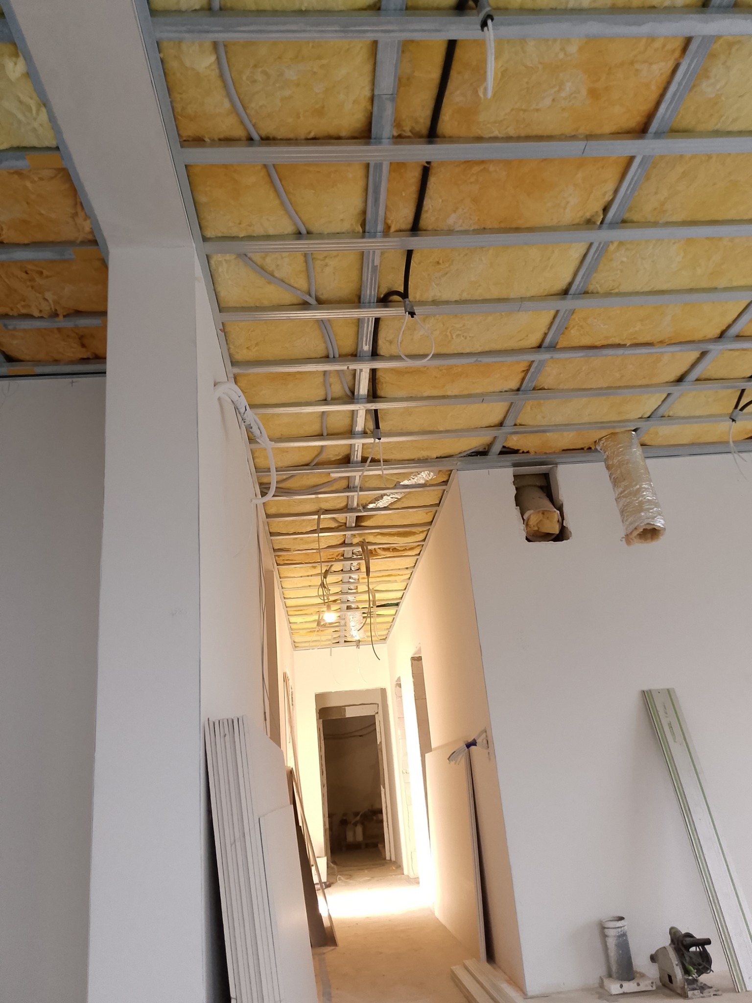 Montaż ocieplenia poddasza z widoczną konstrukcją metalową, wełną mineralną i instalacją elektryczną, z widocznymi płytami karton gips i narzędziami na podłodze.