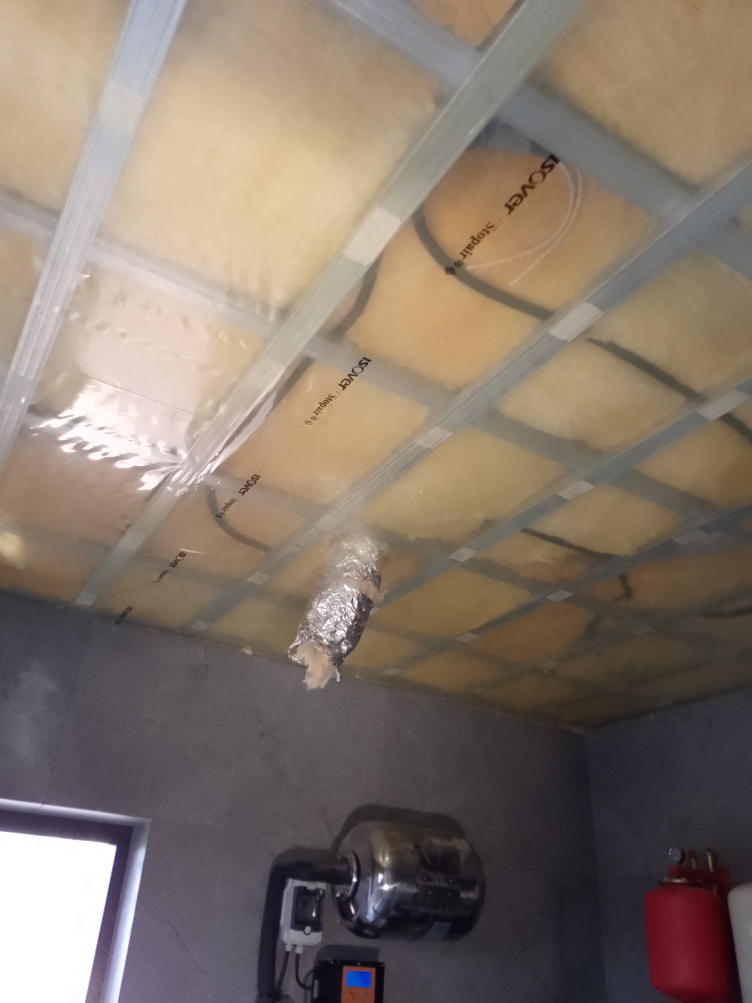 Poddasze z widoczną izolacją z wełny mineralnej Isover, zabezpieczoną folią i metalowym stelażem, fragment instalacji grzewczej przy ścianie.