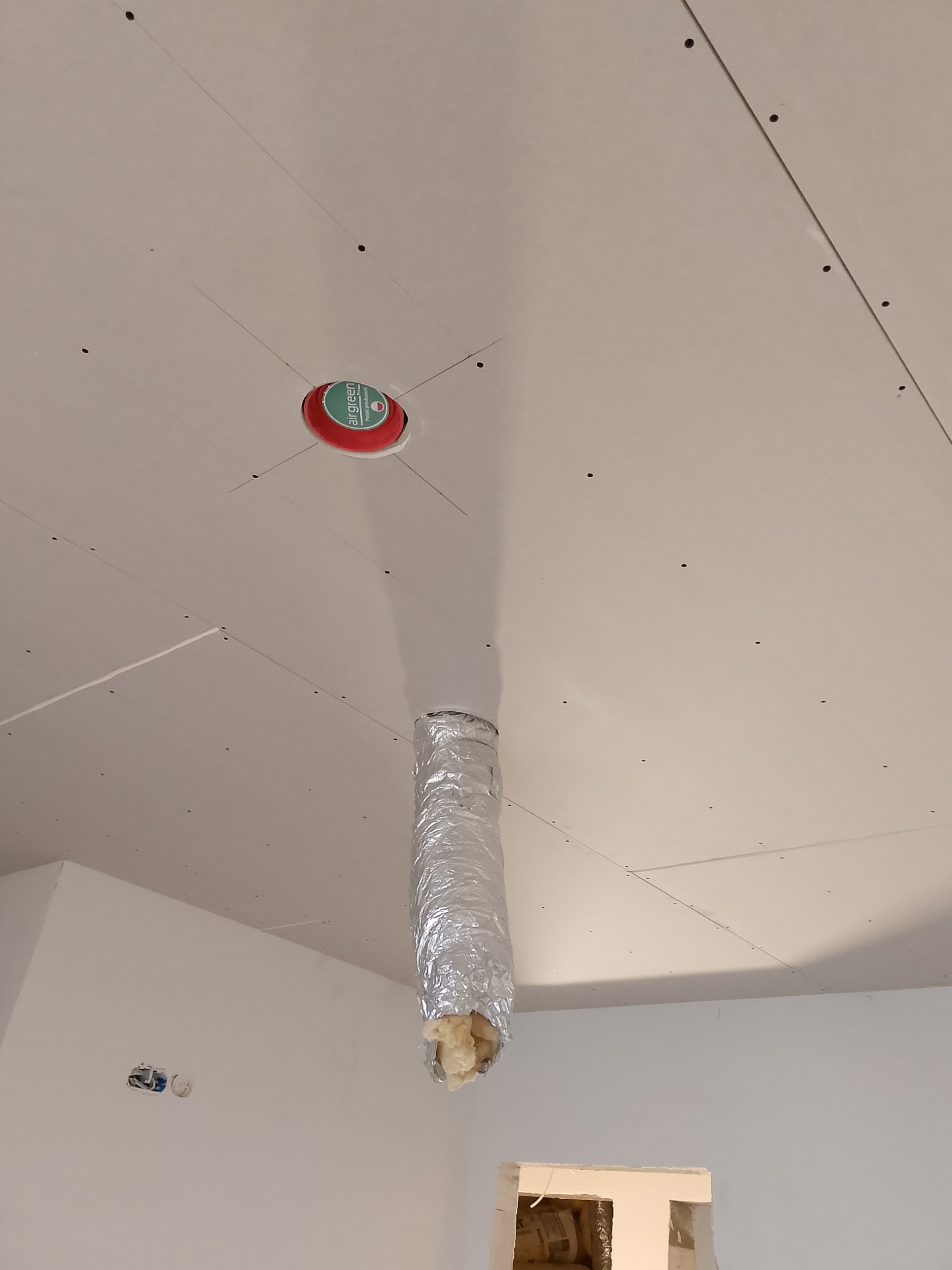 Sufit z płyt kartonowo-gipsowych w trakcie montażu, widoczne otwory na oświetlenie i wentylację, z izolacją wystającą z rury wentylacyjnej, ślady ołówka na łączeniach płyt.