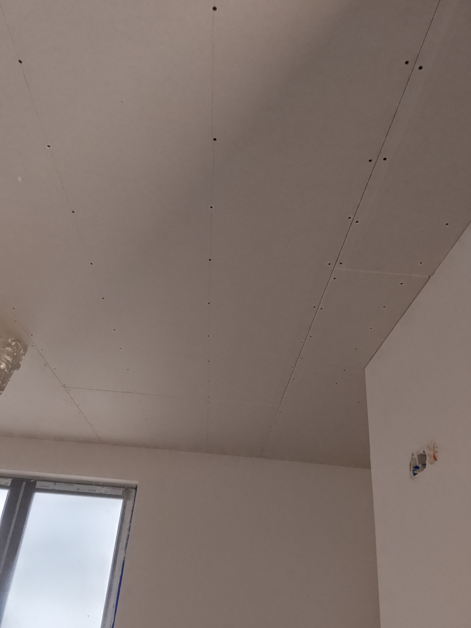 Sufit pokryty płytami gipsowo-kartonowymi, widoczne wkręty i spoiny, przygotowany do dalszych prac wykończeniowych, z oknem w tle.