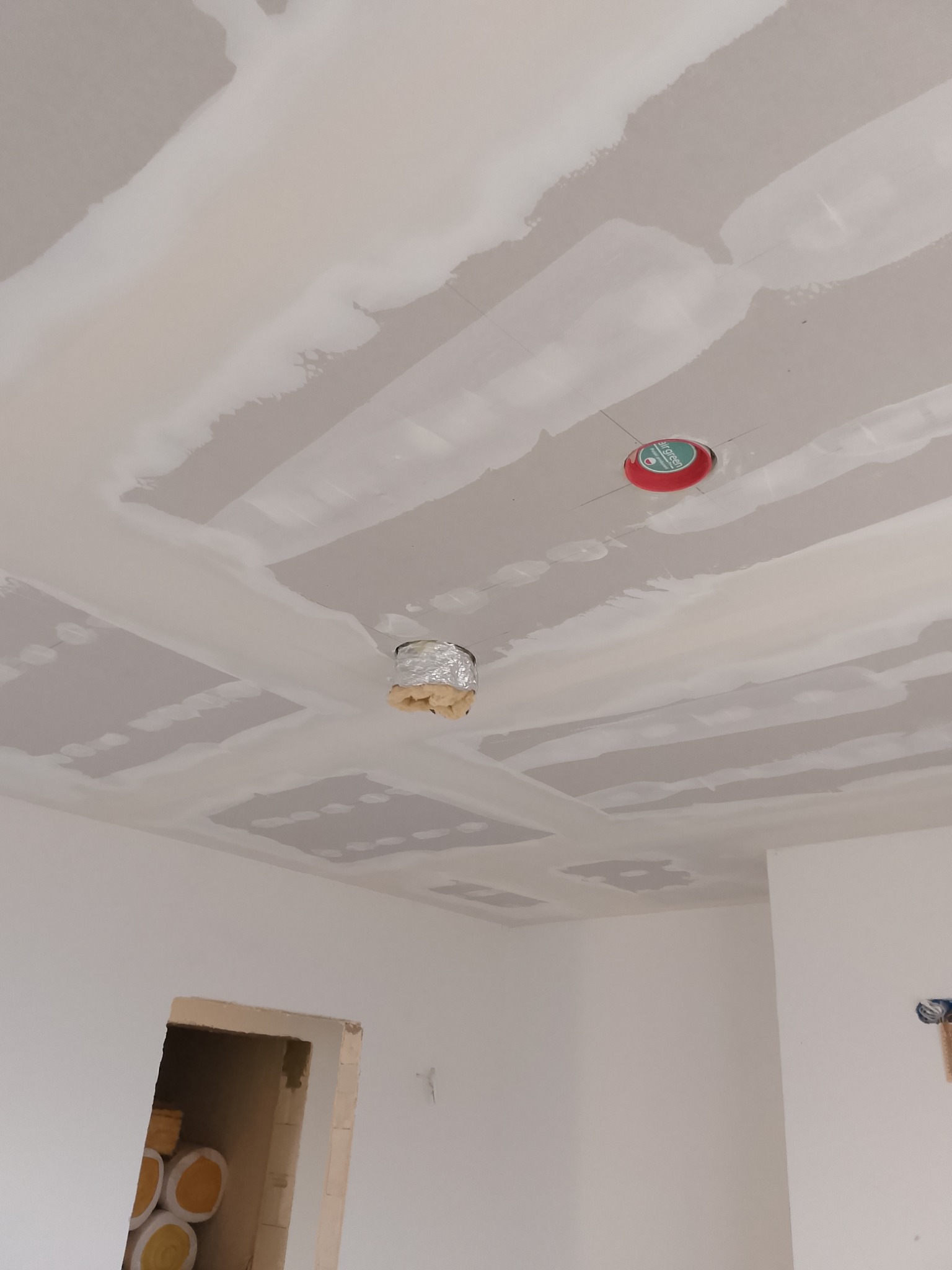 Sufit z płyt gipsowo-kartonowych w trakcie szpachlowania, widoczne łączenia i przygotowane otwory na oświetlenie, jeden z nich zabezpieczony folią aluminiową, a drugi z czerwoną osłoną z napisem.