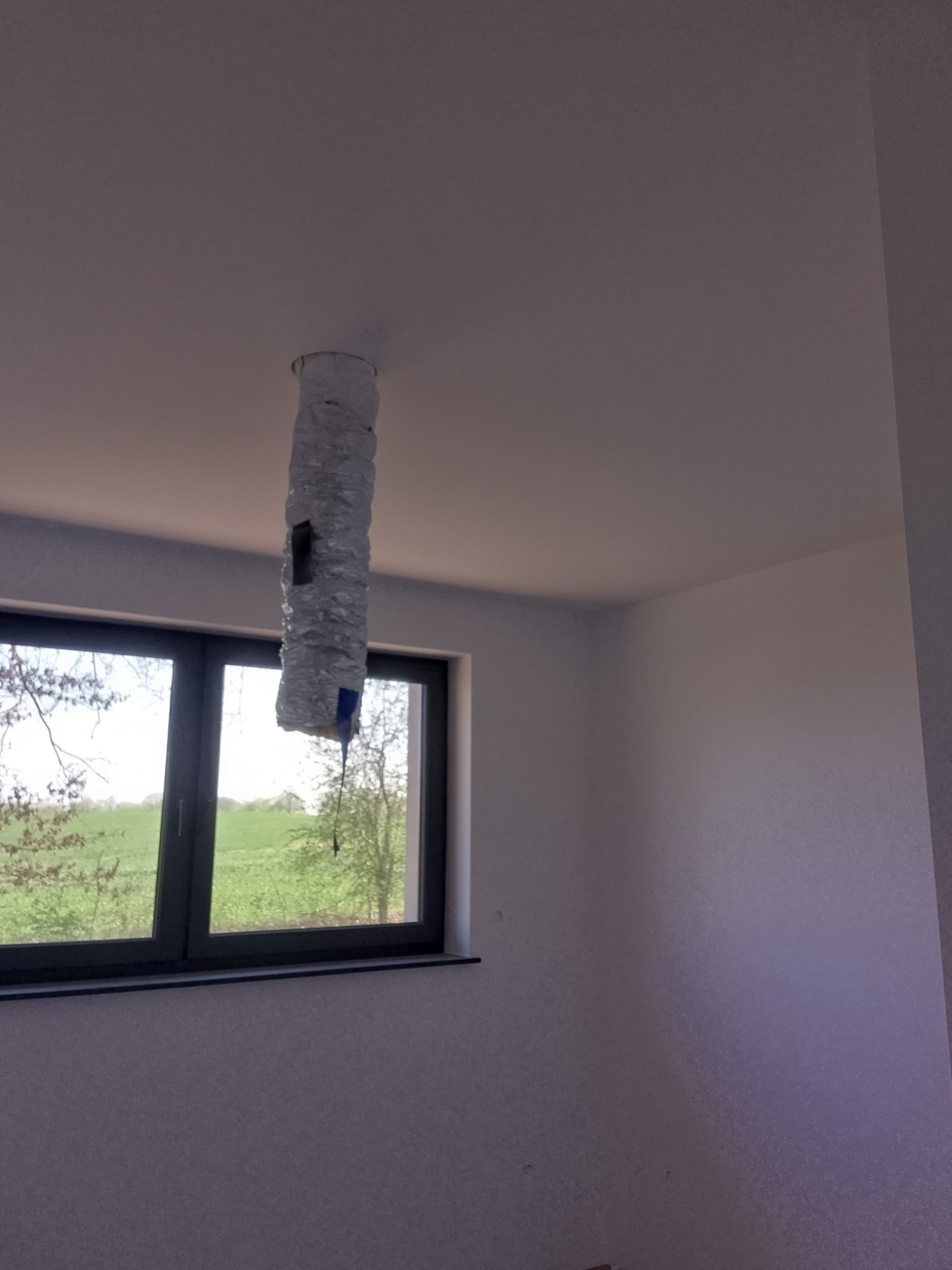 Folia aluminiowa owinięta wokół rury wystającej z sufitu w jasnym pokoju z oknem. Prace przygotowawcze do montażu oświetlenia.