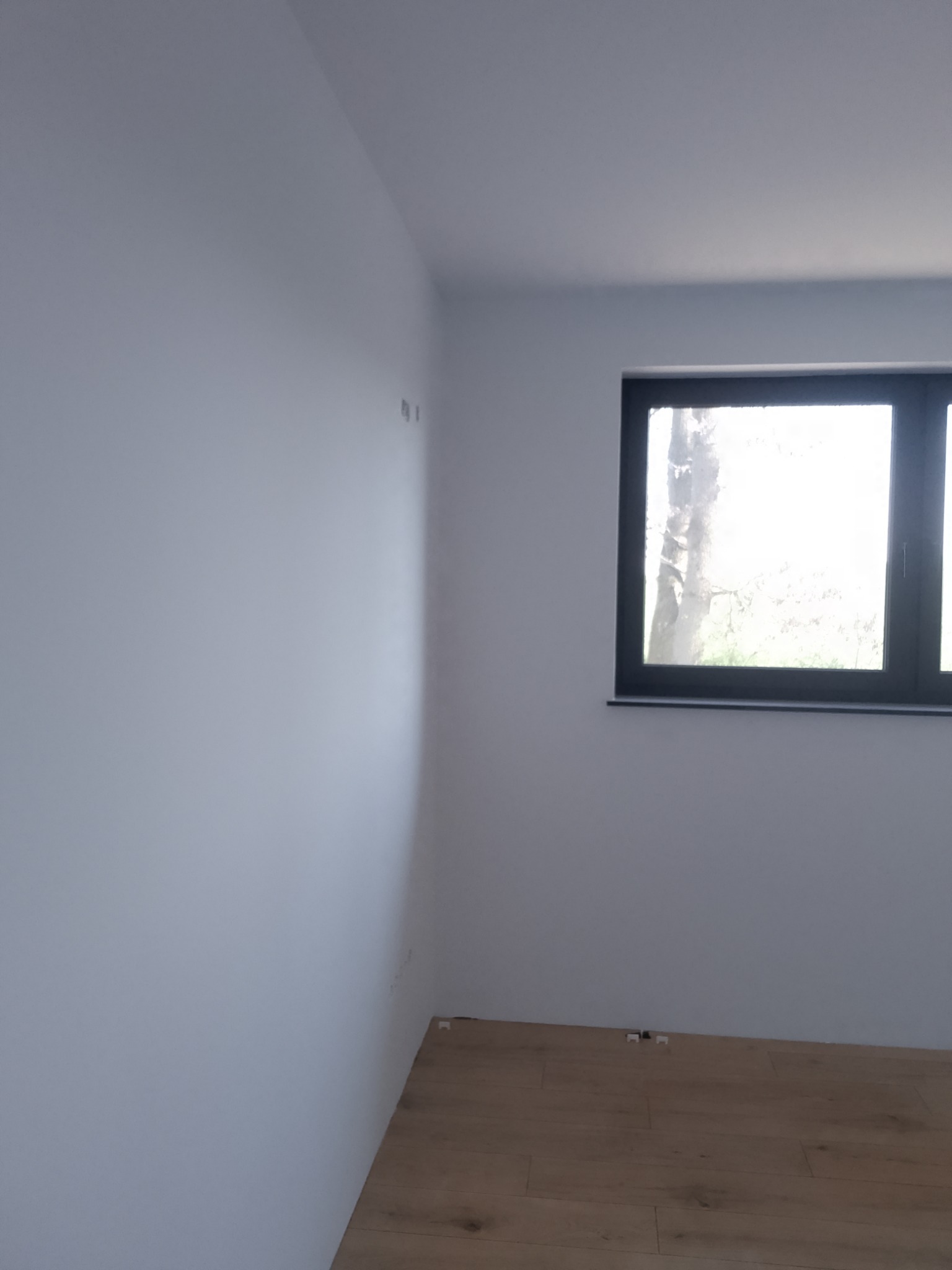 Jasne, pomalowane na biało wnętrze z drewnianą podłogą i oknem z czarną ramą, ukazujące minimalistyczny styl wykończenia.