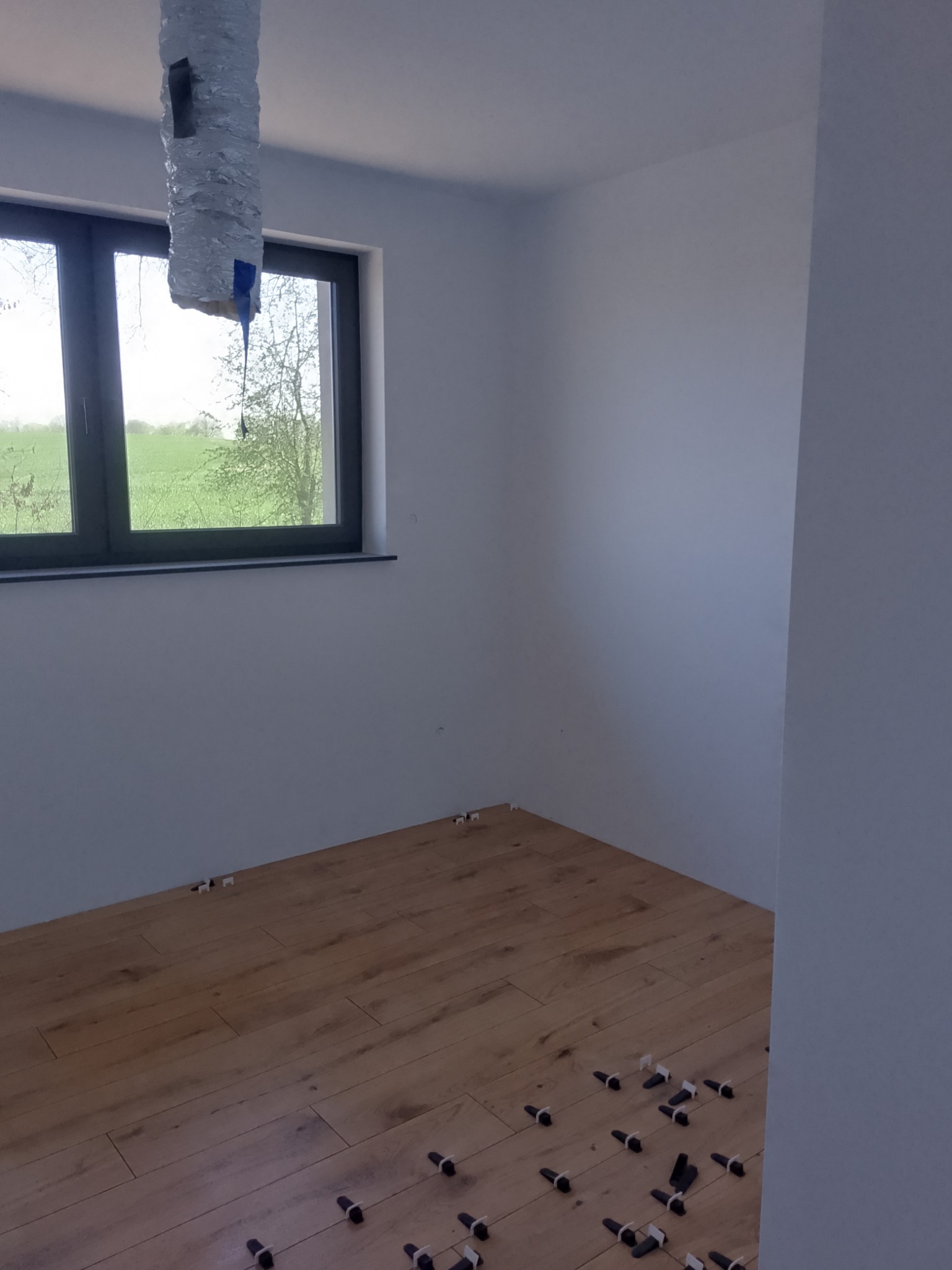 Jasne panele podłogowe w trakcie układania, widoczne kliny dystansowe, okno z widokiem na zieleń. Białe ściany i rura wentylacyjna pod sufitem.