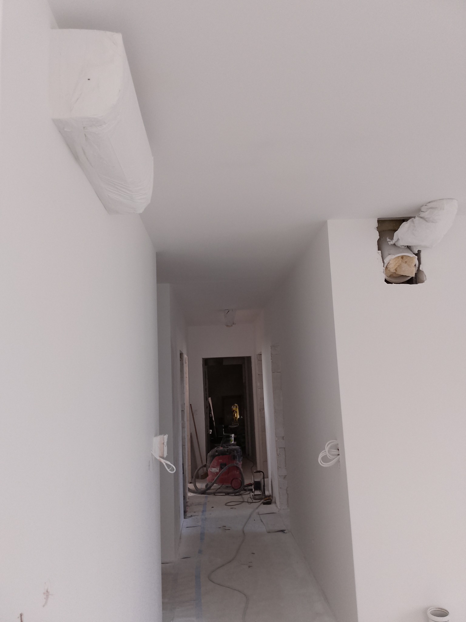 Wnętrze w trakcie prac wykończeniowych: białe ściany, zabezpieczone elementy (klimatyzator, rury), widoczny sprzęt budowlany w głębi korytarza. Minimalistyczny, surowy styl.