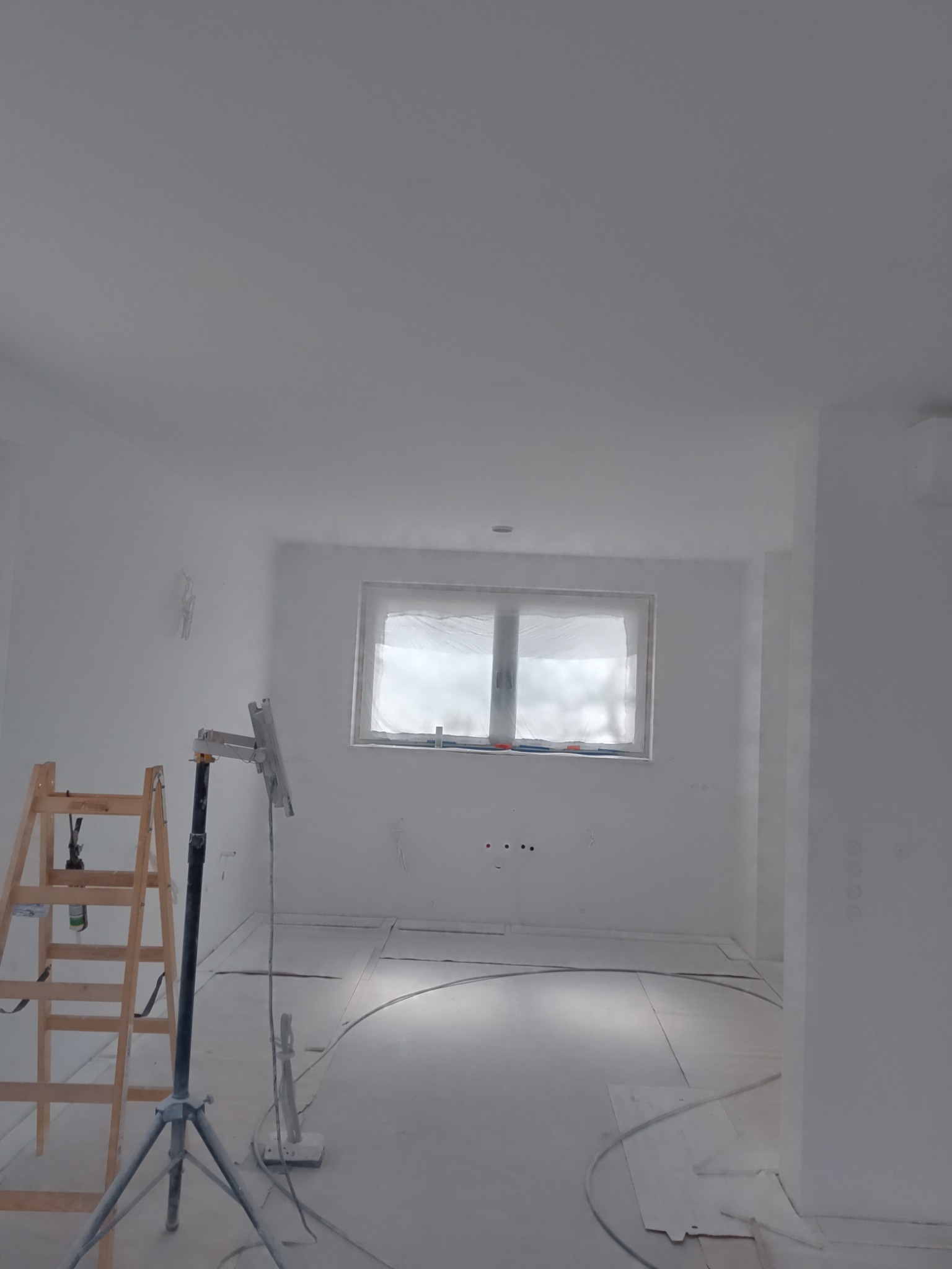 Białe, świeżo pomalowane wnętrze z drabiną i osłoniętym oknem. Widoczny sprzęt malarski sugeruje prace wykończeniowe w trakcie realizacji.