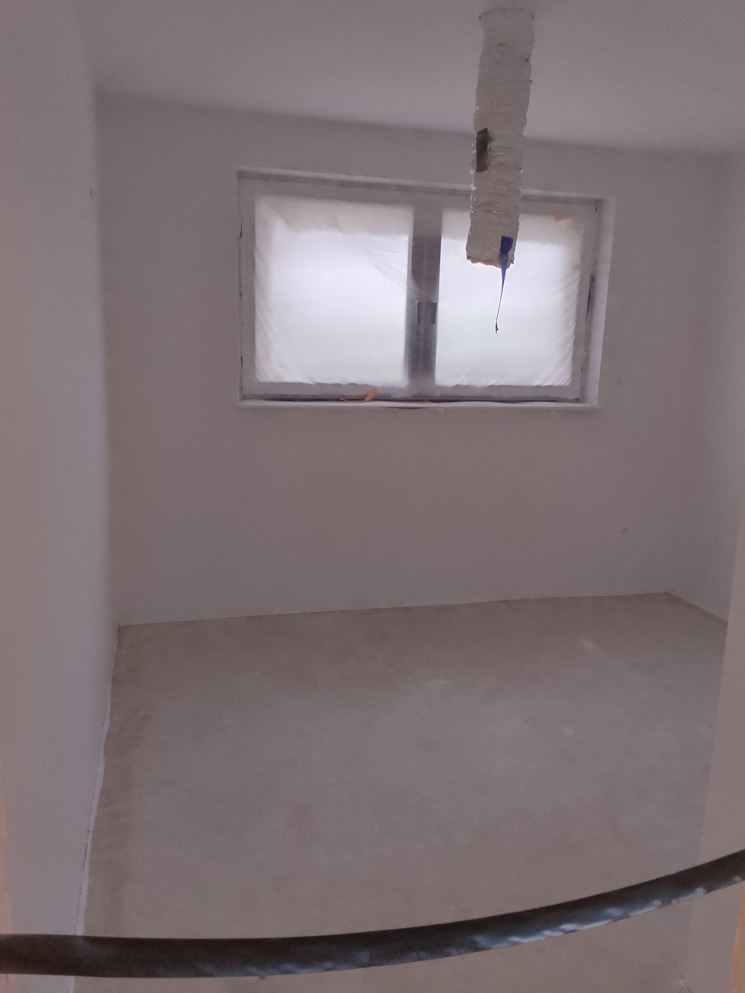 Pokój po wylewce podłogi, z oknem zabezpieczonym folią i rurą owiniętą taśmą. Ściany przygotowane do dalszych prac. Widoczne ślady farby.