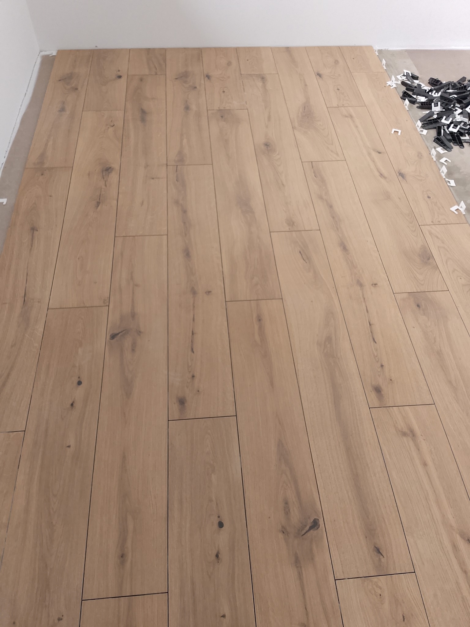 Fragment układania paneli podłogowych z widocznymi elementami dystansowymi przy ścianie, ukazujący postęp prac. Jasne panele z wyraźnym wzorem drewna.