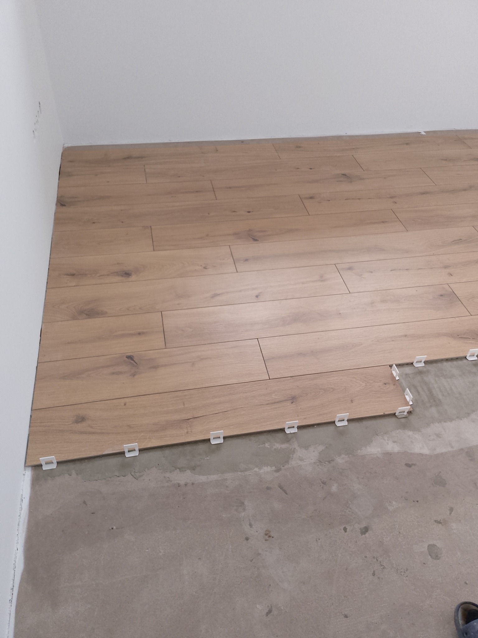 Układanie jasnych paneli podłogowych, widoczne klipsy dystansowe przy ścianie. Częściowo wykończona podłoga na surowym podłożu. Ujęcie z góry.