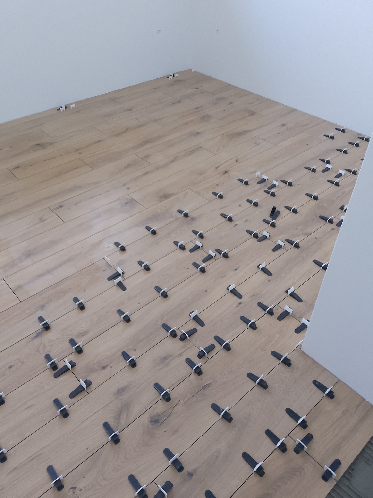 Układanie paneli podłogowych z użyciem klipsów poziomujących w jasnym wnętrzu. Widoczny fragment ściany i podłogi z panelami w odcieniach drewna.