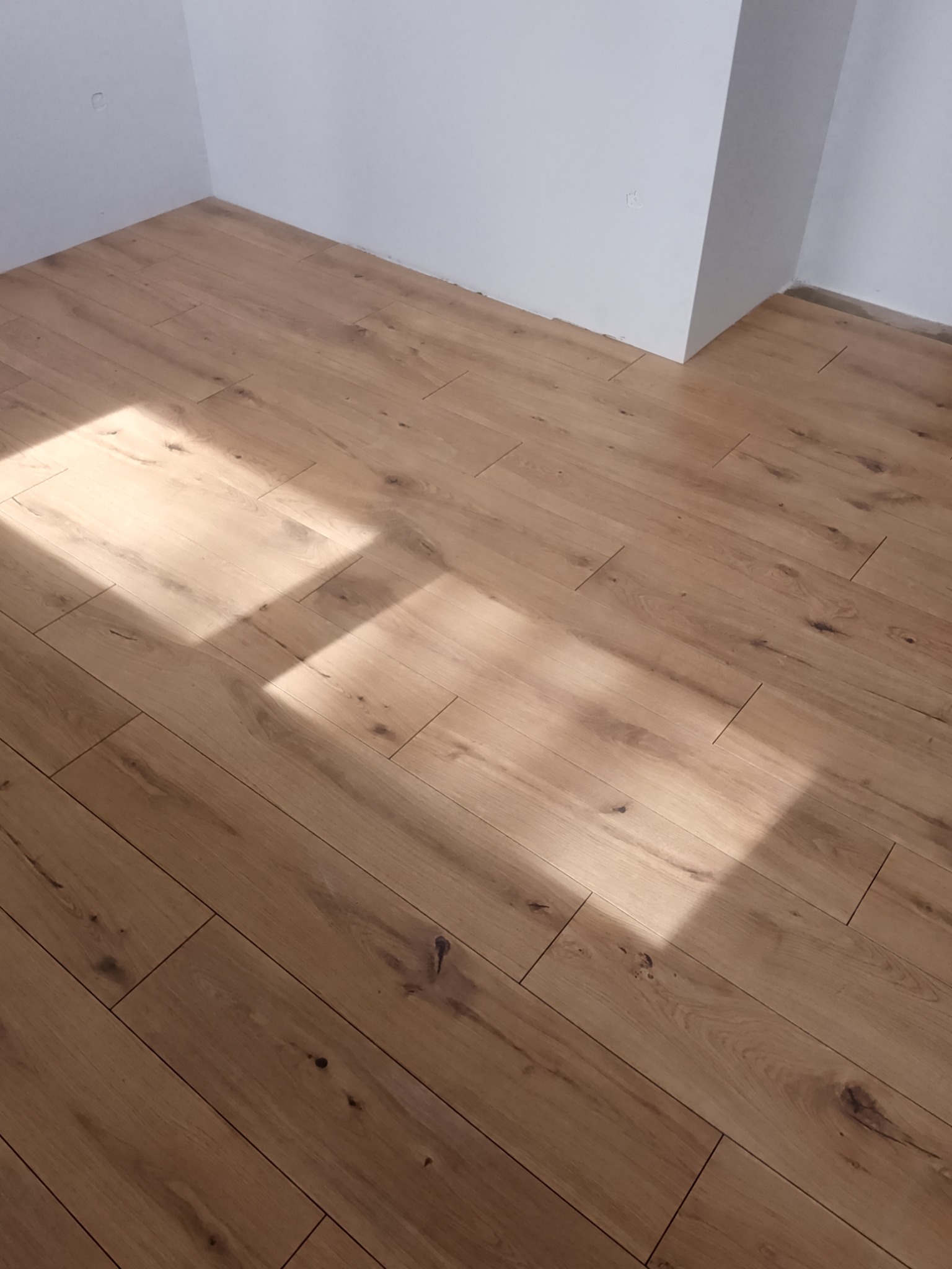 Ułożone panele podłogowe w jasnym odcieniu drewna, ukośnie ułożone względem kadru, z wyraźnym wzorem słojów i odbiciami światła słonecznego w rogu pomieszczenia.