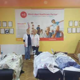 Wnętrze gabinetu z leżącymi osobami na łóżkach rehabilitacyjnych Ceragem, personel medyczny stojący przed plakatem reklamowym, ściany ozdobione dyplomami.