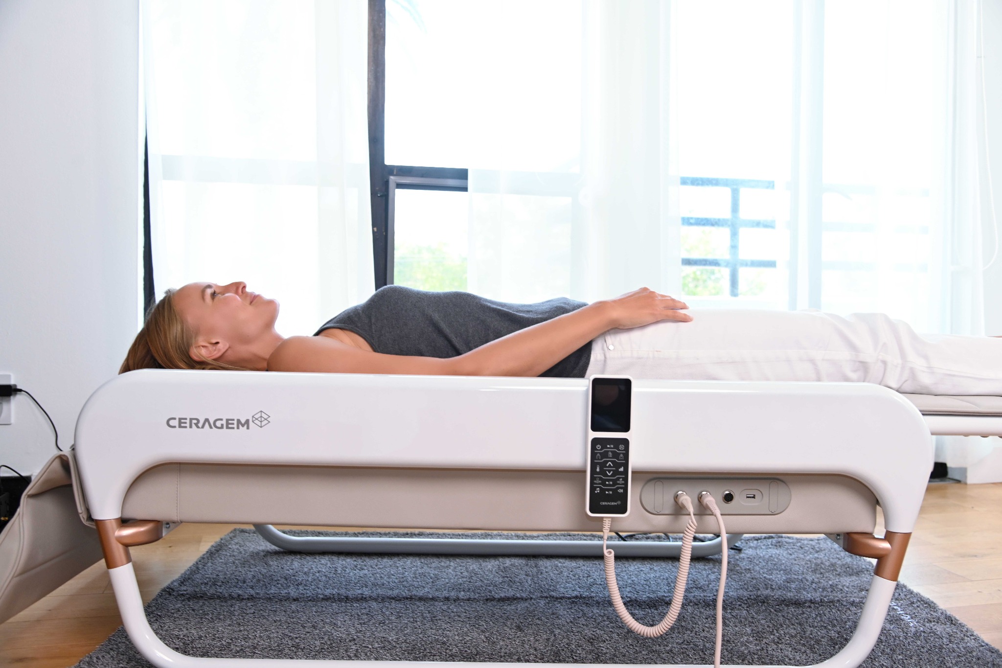 Kobieta relaksuje się na automatycznym łóżku do masażu Ceragem w jasnym wnętrzu, z pilotem sterującym i widocznymi elementami konstrukcyjnymi.