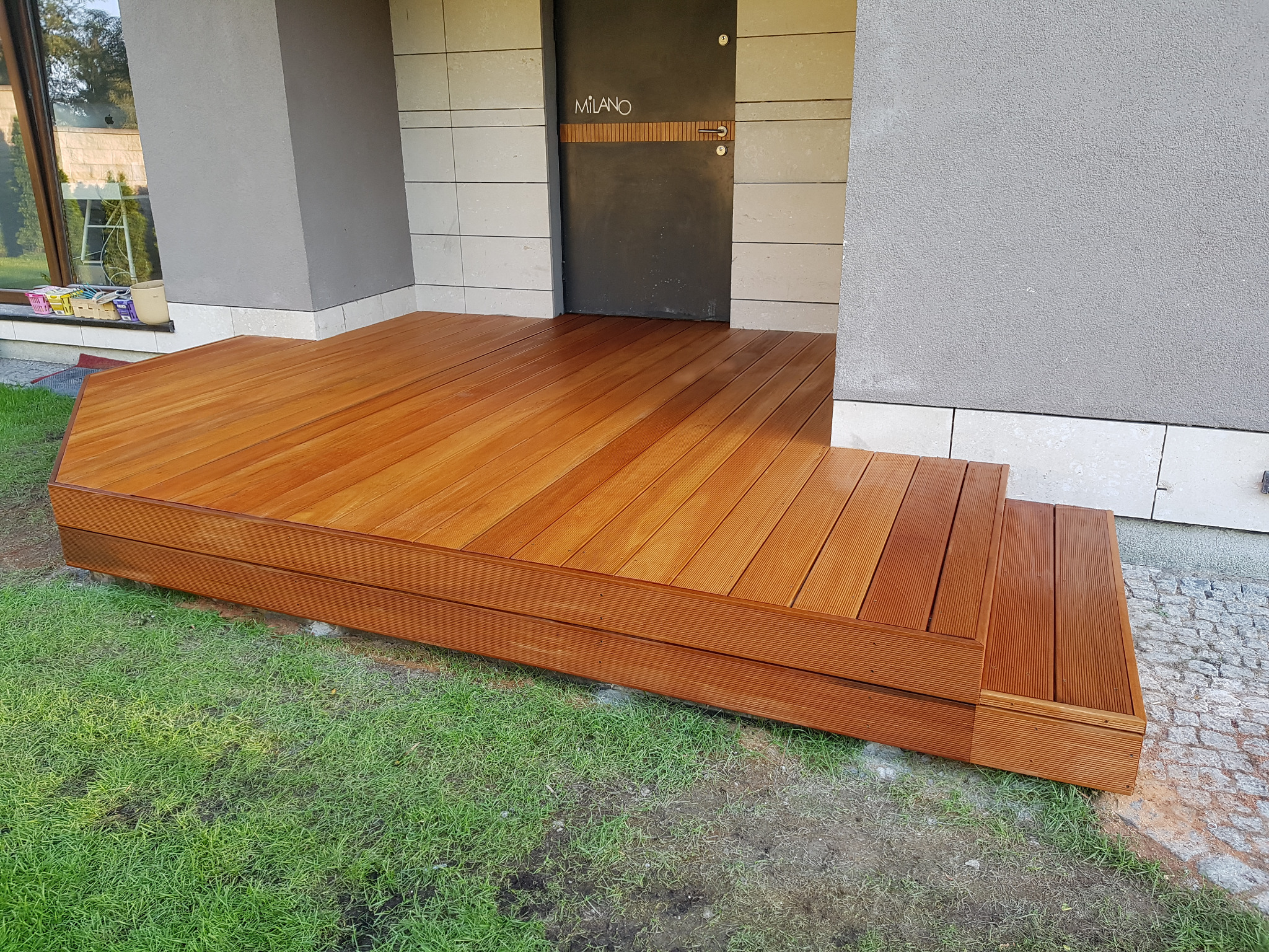 Nowoczesny drewniany taras z trzema stopniami prowadzący do drzwi wejściowych domu, z widoczną zieloną trawą i szarą elewacją.