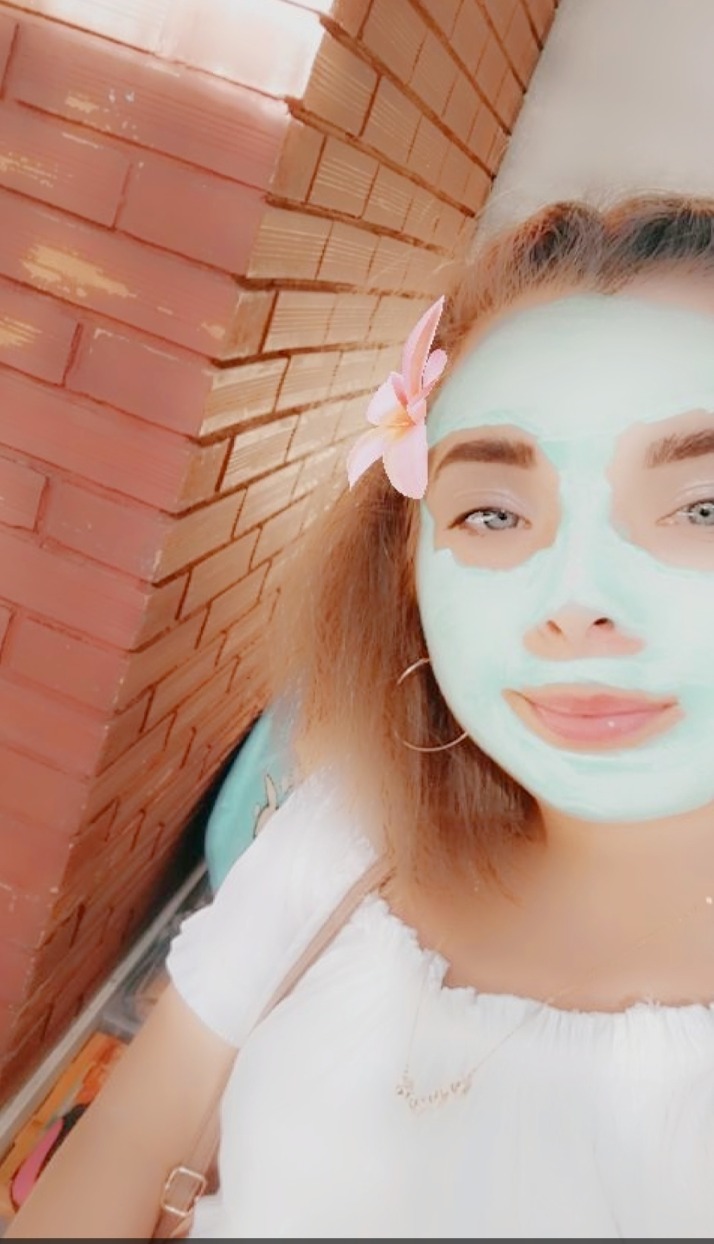 Portret kobiety z zieloną maseczką na twarzy i różowym kwiatem we włosach, uśmiechnięta, z widoczną częścią ceglanej ściany w tle.