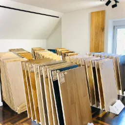 Ekspozycja różnorodnych paneli i desek podłogowych w salonie, ułożonych w rzędach na stojakach, z widocznym wzorem parkietu na podłodze i oświetleniem punktowym.