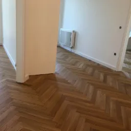 Nowo ułożona, lakierowana podłoga drewniana w jodełkę, widok z korytarza na jasne, odnowione wnętrze z białymi ścianami i grzejnikiem.