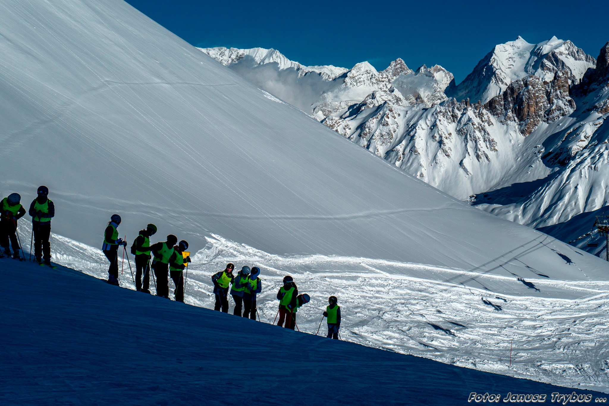 Grupa narciarzy w kaskach i jaskrawych kamizelkach na tle ośnieżonych gór, widok z dołu.
