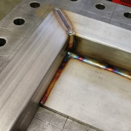 IRONFUN Kamil Banaszewski - Metalowa rama połączona spoiną, widoczne tęczowe przebarwienia termiczne na spawie, ułożona na perforowanym stole spawalniczym.