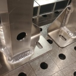 IRONFUN Kamil Banaszewski - Dwa spawane, aluminiowe wsporniki kątowe z otworami, leżące na metalowym stole spawalniczym z siatką otworów, widoczne ślady obróbki.