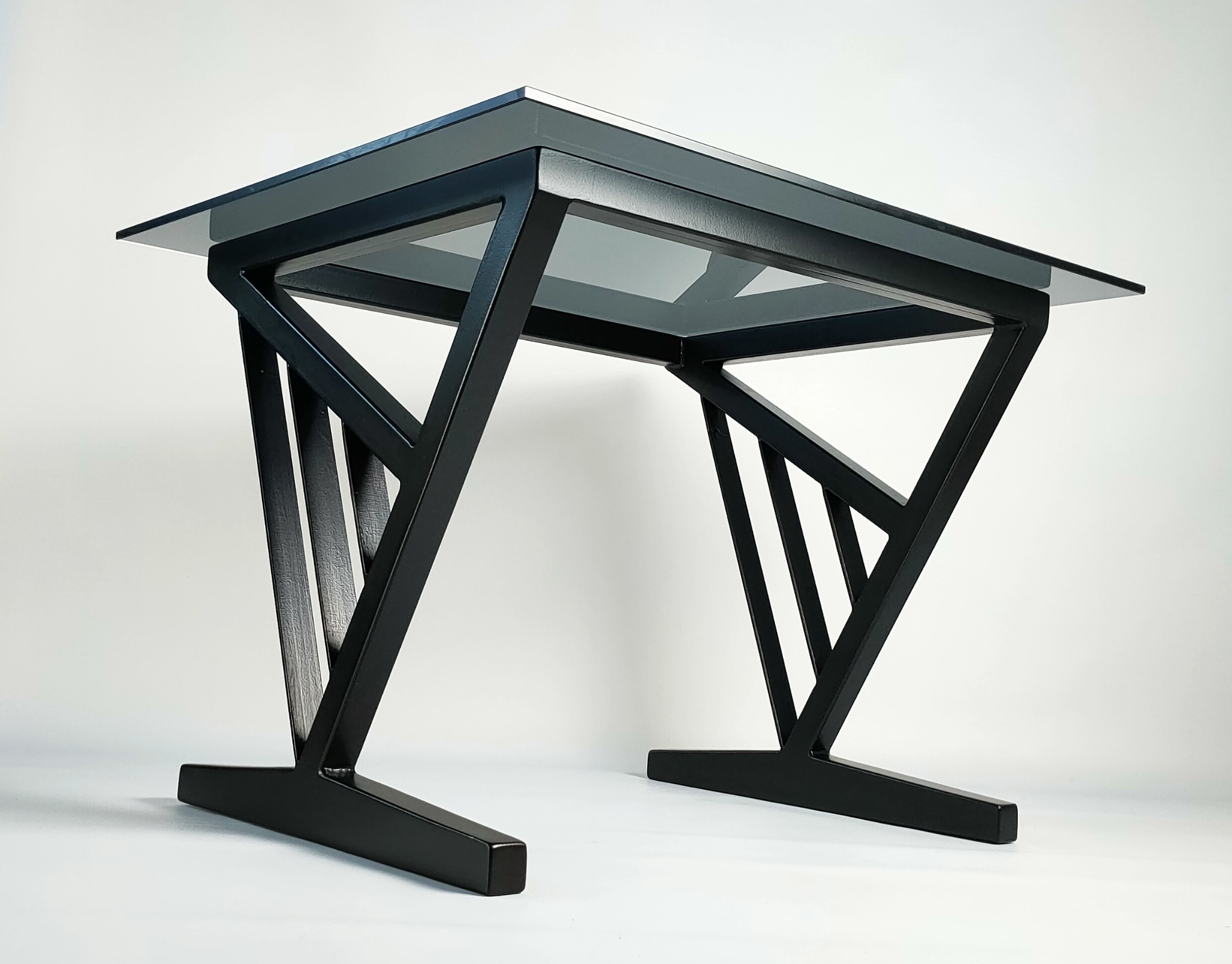 Nowoczesny stolik kawowy z czarną, metalową konstrukcją i szklanym blatem, ustawiony na białym tle, prezentujący geometryczny design nóg.