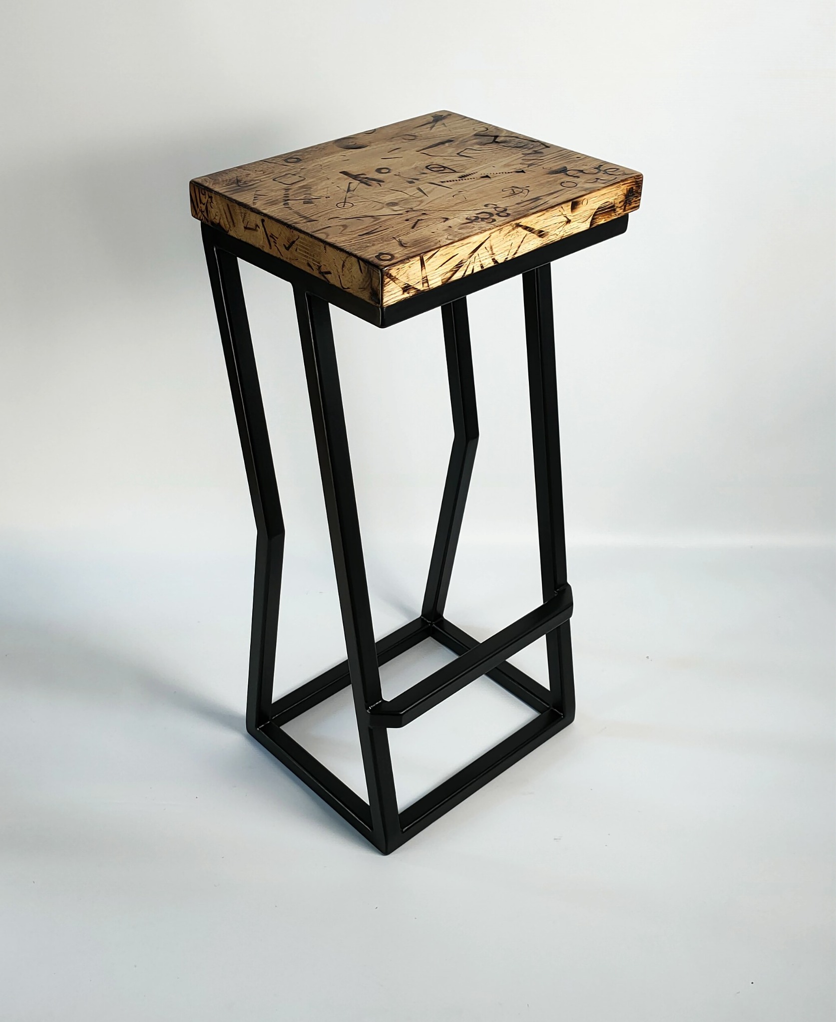 Wysoki stołek barowy z czarną, metalową ramą o geometrycznym kształcie i drewnianym siedziskiem z wygrawerowanymi wzorami, prezentowany na białym tle.