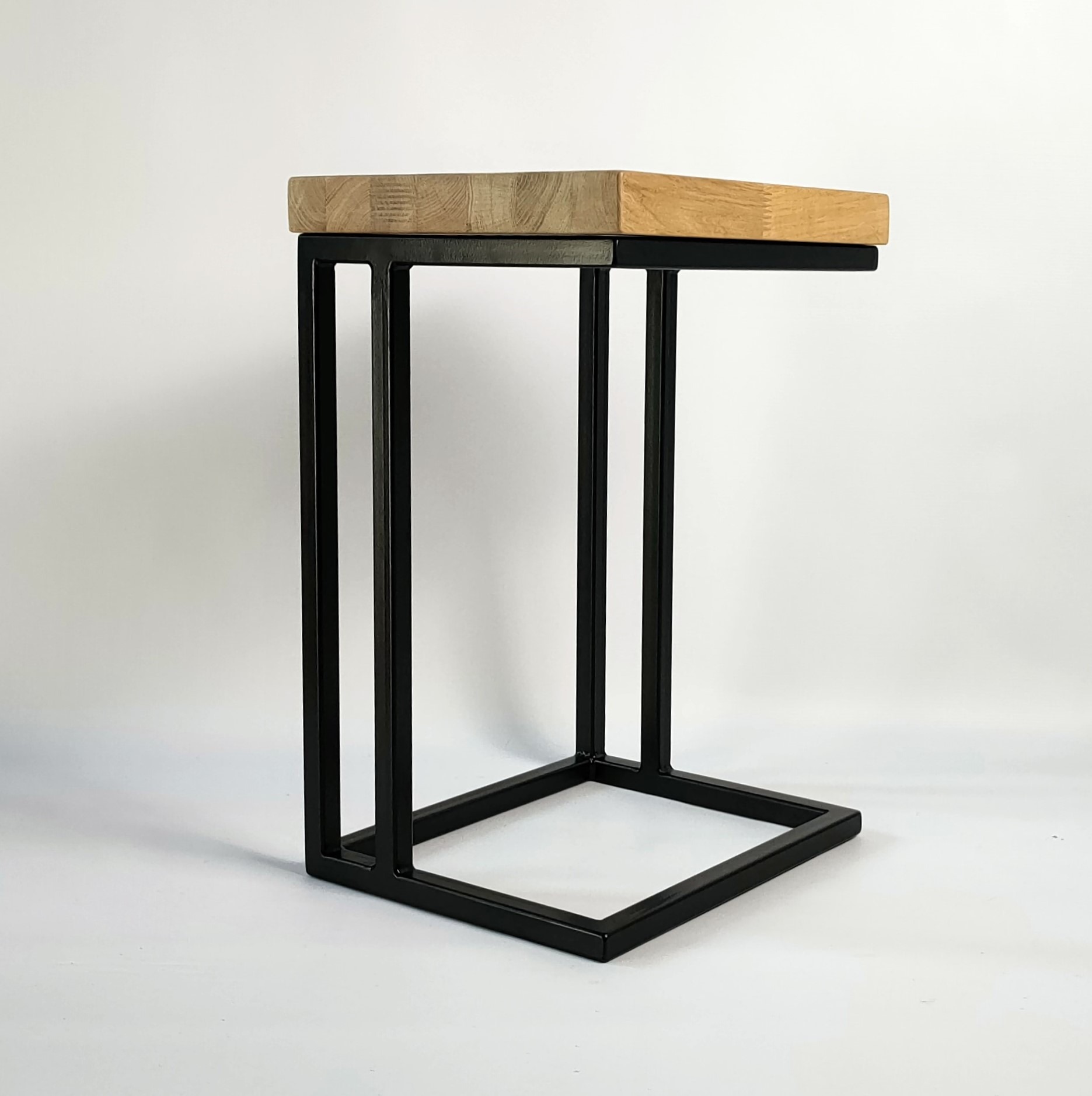 Stolik pomocniczy z blatem z jasnego drewna i czarną, metalową podstawą w kształcie litery C, na białym tle.