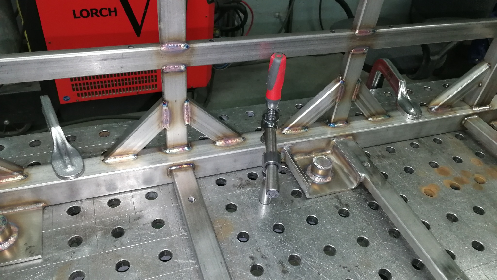 Metalowa konstrukcja spawana na perforowanym stole spawalniczym, z widocznymi śladami spawania i narzędziami mocującymi, w tle czerwony agregat spawalniczy marki Lorch.