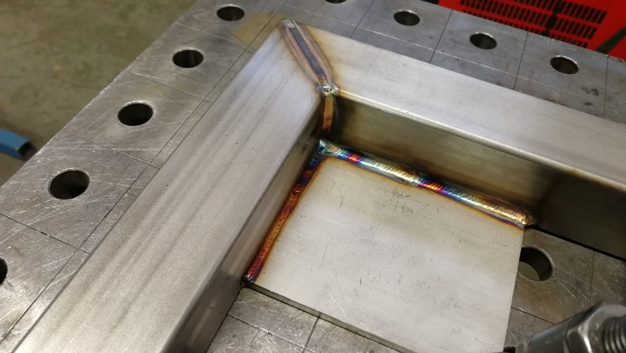 Metalowa rama połączona spoiną, widoczne tęczowe przebarwienia termiczne na spawie, ułożona na perforowanym stole spawalniczym.