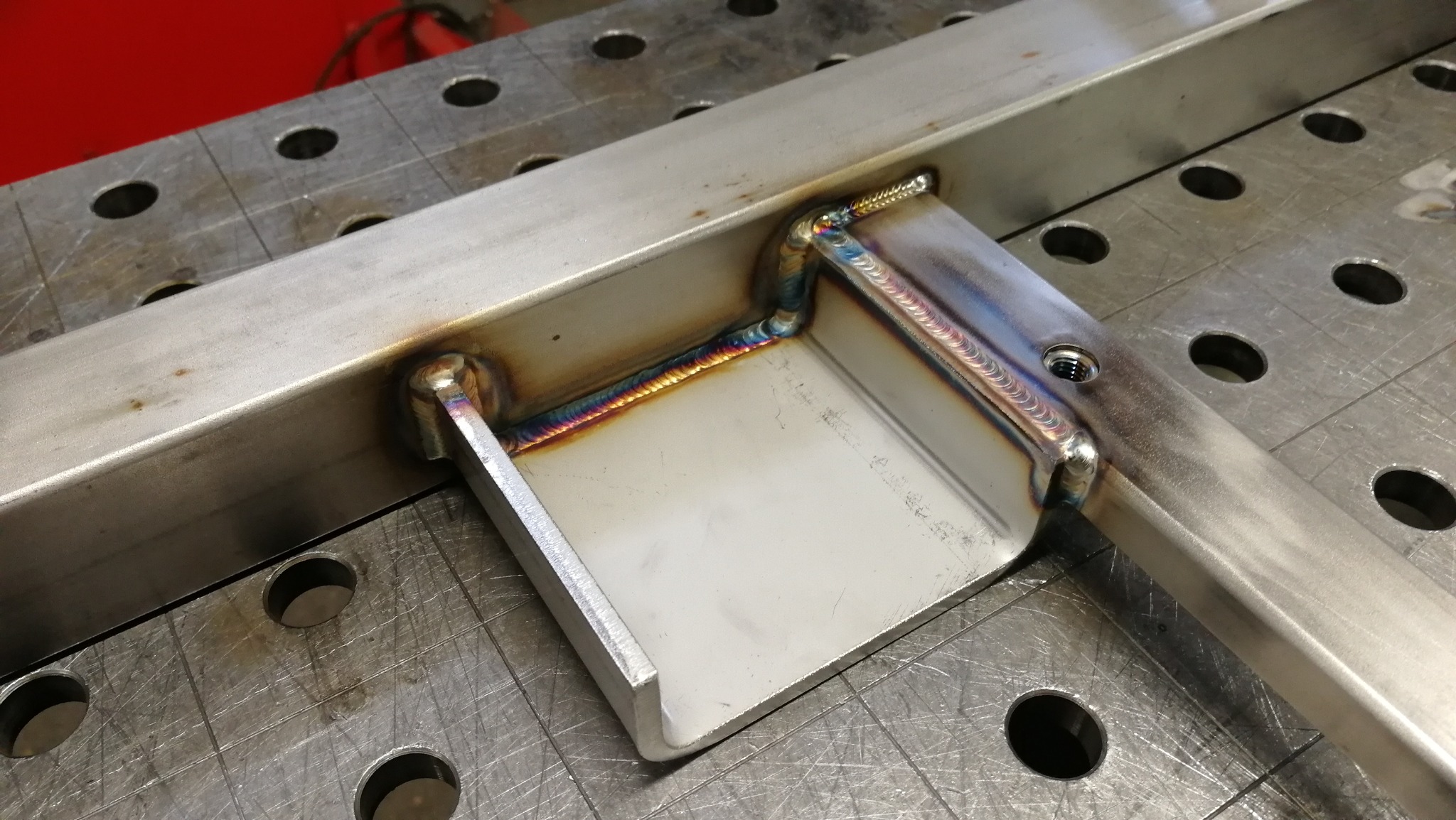 Szlifowany metalowy profil spawany do elementu konstrukcyjnego na perforowanym stole spawalniczym, widoczne ślady ciepła po spawaniu i gwintowany otwór.