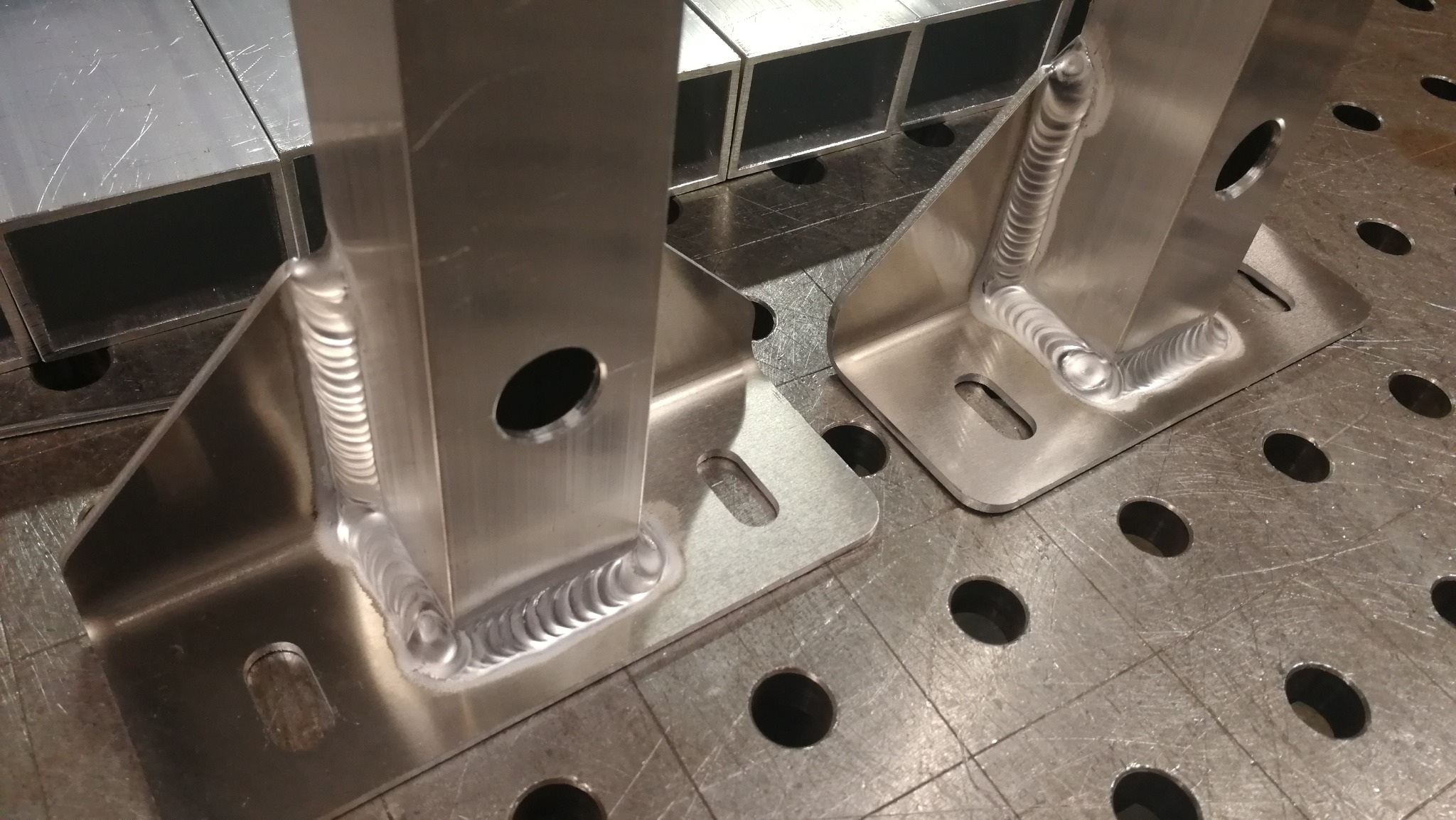 Dwa spawane, aluminiowe wsporniki kątowe z otworami, leżące na metalowym stole spawalniczym z siatką otworów, widoczne ślady obróbki.