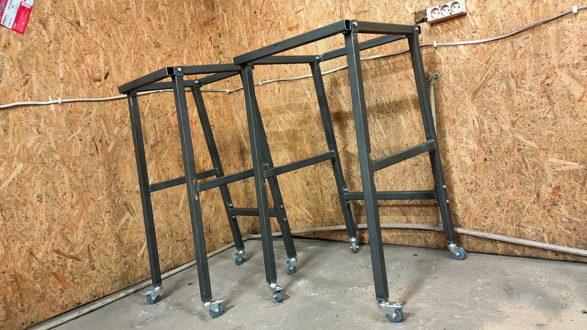 Dwie surowe, metalowe konstrukcje na kółkach, prawdopodobnie w warsztacie, z widocznymi spawami i śrubami, ustawione na tle ściany z płyty OSB.