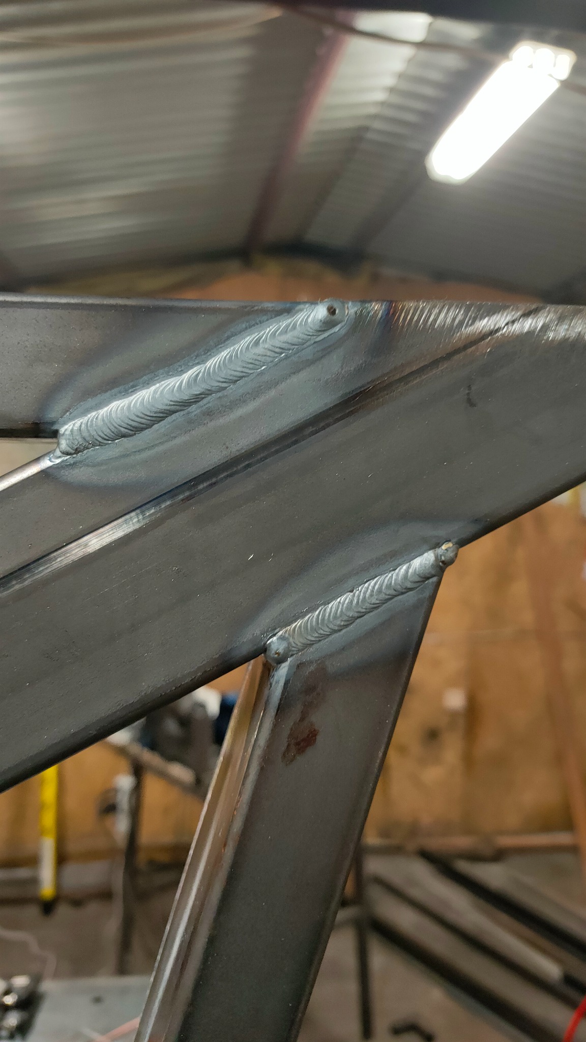 Szczegółowe zbliżenie na spoiny łączące elementy metalowej konstrukcji, widoczne regularne ślady spawania na szarym metalu w warsztatowym otoczeniu.