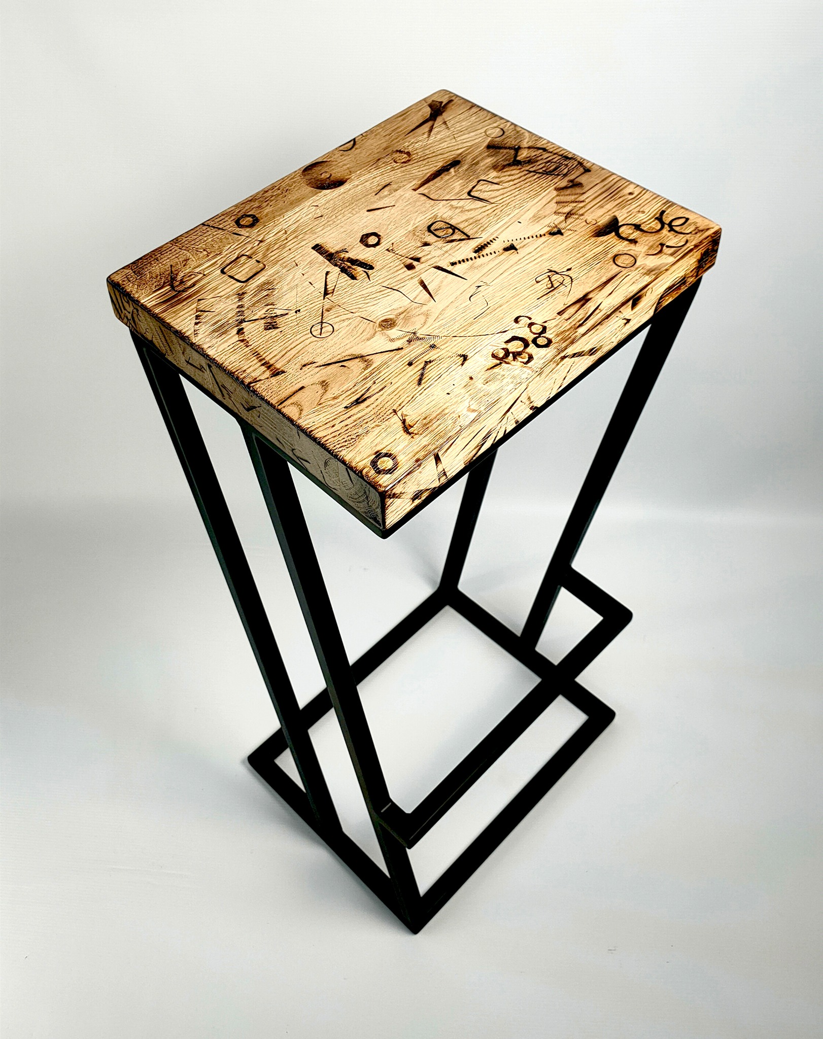 Stolik barowy z blatem z jasnego drewna zdobionego wypalanymi wzorami i geometryczną podstawą z czarnych, metalowych prętów.