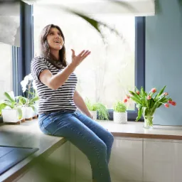 Kobieta w bluzce w paski siedzi na blacie kuchennym, podrzucając pomarańczę w powietrze, w tle okno z widokiem na drzewa i kwiaty w wazonach.