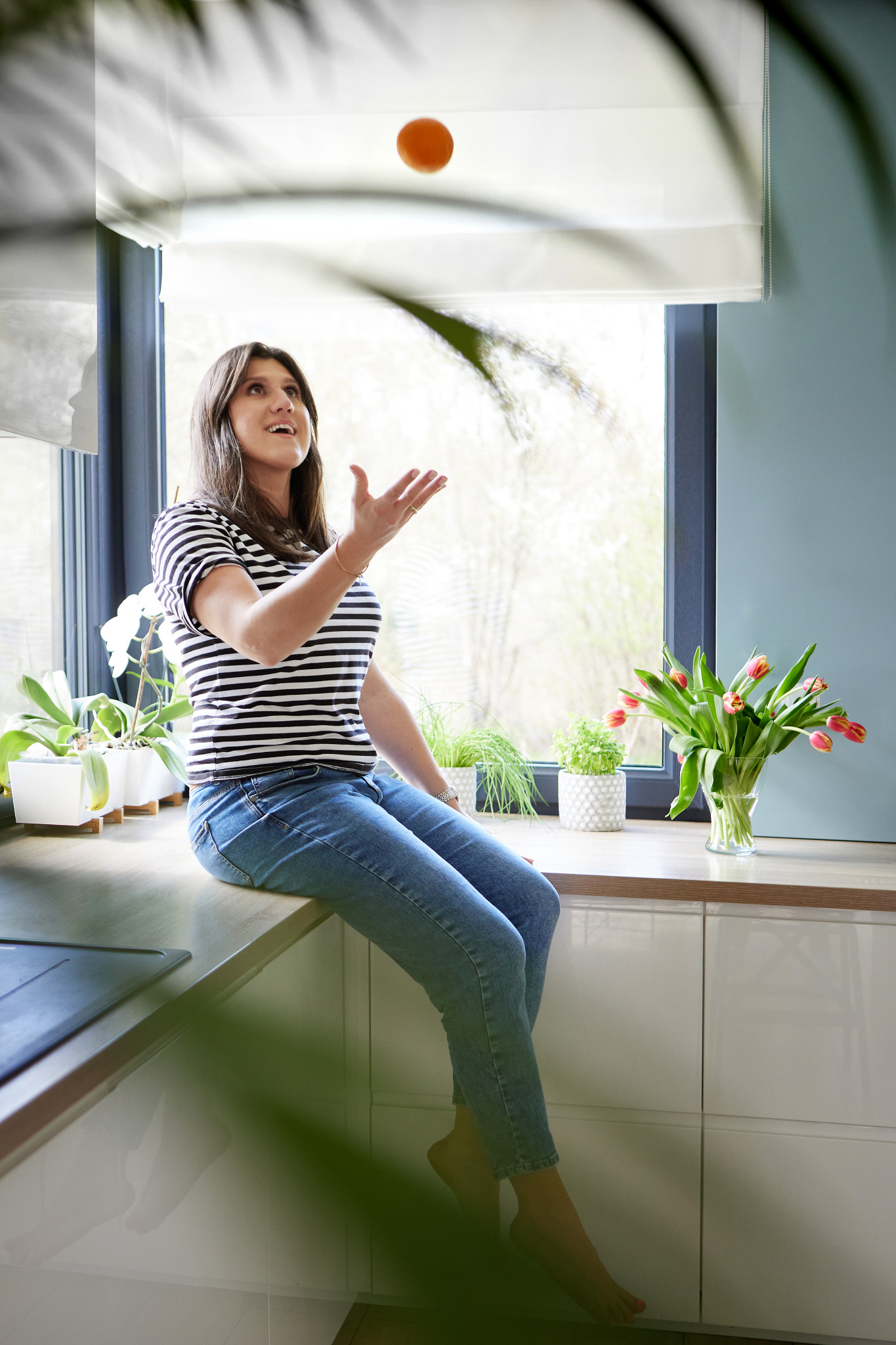 Kobieta w bluzce w paski siedzi na blacie kuchennym, podrzucając pomarańczę w powietrze, w tle okno z widokiem na drzewa i kwiaty w wazonach.