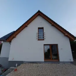 Dwukondygnacyjny dom z białymi ścianami i brązowymi ramami okiennymi, widok z zewnątrz na stertę żwiru przed budynkiem.