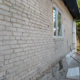 Ceglana ściana budynku przed pracami elewacyjnymi, widoczne ubytki w dolnej części, okno z uszczelnieniami i przygotowane płyty styropianowe do ocieplenia.