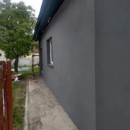 Świeżo otynkowana i pomalowana na szaro elewacja budynku jednorodzinnego, widok z boku z fragmentem chodnika i ogrodzenia.