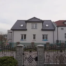 Dom jednorodzinny z szarą dachówką i jasną elewacją, widoczny zza ozdobnego, metalowego ogrodzenia z kamiennymi słupkami. Okna zabezpieczone niebieską folią malarską, z tyłu widoczna drabina oparta...