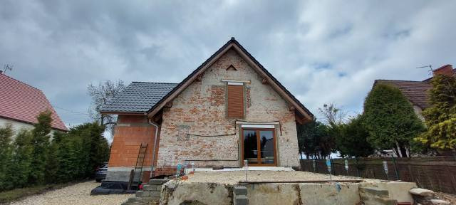 Ceglany budynek w trakcie prac: widoczna nowa stolarka okienna, częściowo otynkowana ściana z odsłoniętą cegłą, nowa dachówka, rusztowanie i drabina oparta o ścianę z pustaków.