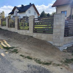 METAL-MAT Mateusz Kasprzyk - Nowoczesne ogrodzenie z betonowych bloczków i poziomych, brązowych przęseł, z widocznym fragmentem bramy wjazdowej, na tle dwóch domów jednorodzinnych w słoneczny dzień.