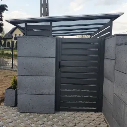 Nowoczesna, pozioma brama wjazdowa w kolorze grafitowym z zadaszeniem z przezroczystego tworzywa, osadzona na betonowych słupkach i kostce ażurowej wypełnionej kamieniami. Obok donica z małą...
