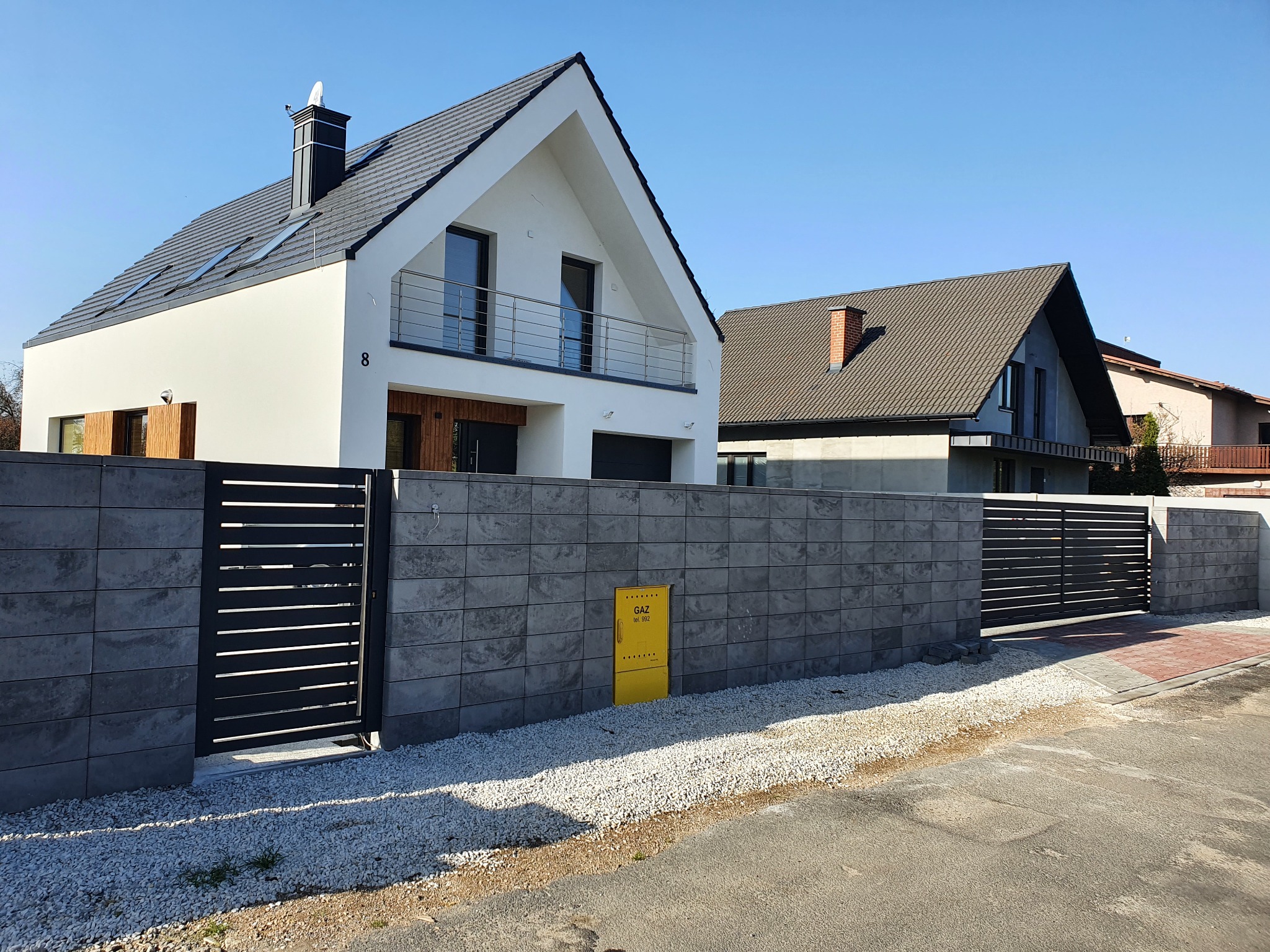 Nowoczesna posesja z szarego betonu z poziomymi, czarnymi przęsłami ogrodzeniowymi i bramą wjazdową, na tle białego domu z ciemną dachówką i błękitnego nieba.