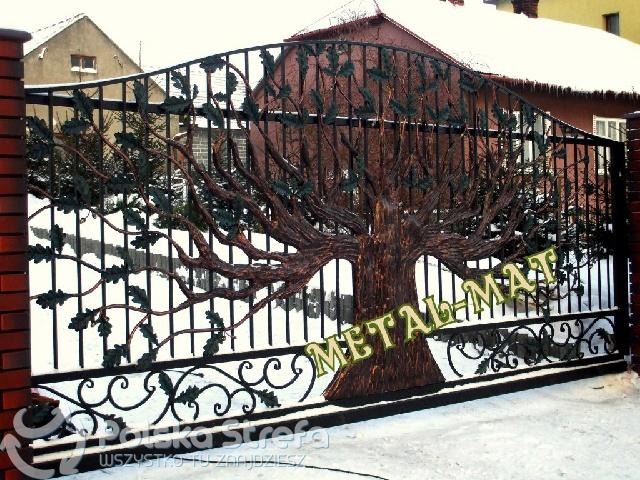 Kuta brama wjazdowa z motywem drzewa dębu, czarne metalowe pręty i liście na tle ośnieżonego podjazdu i ceglanych słupków.