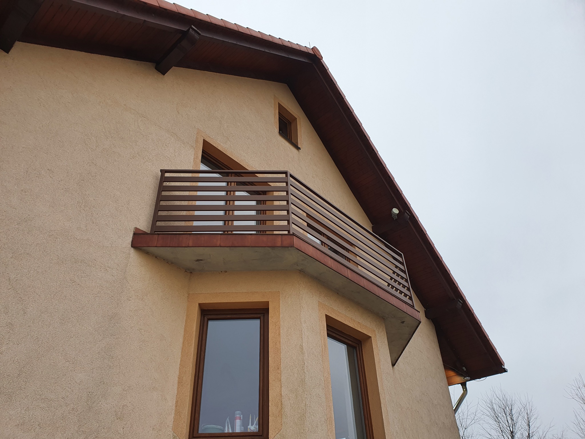 Zewnętrzny widok domu z beżową elewacją, brązowym dachem i balkonem z metalową balustradą o poziomym układzie szczebli.