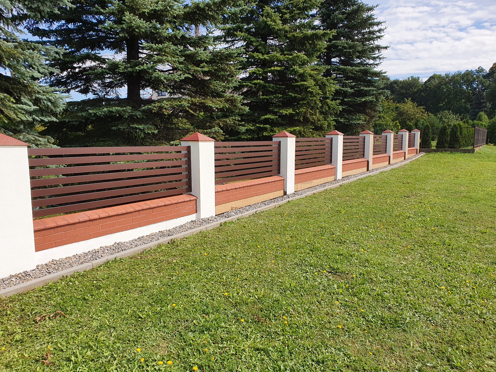 Nowoczesne ogrodzenie panelowe z brązowymi przęsłami, białymi słupkami zwieńczonymi ceglastymi daszkami i cokołem z cegły klinkierowej, otoczone zielonym trawnikiem i wysokimi drzewami iglastymi.